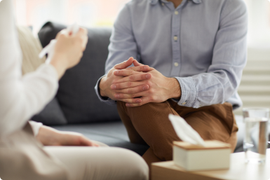 Pessoa sentada em um ambiente acolhedor, com as mãos cruzadas sobre os joelhos, em uma conversa atenta com outra pessoa segurando um lenço. Há uma caixa de lenços sobre a mesa, sugerindo um cenário de escuta e acolhimento, possivelmente em uma sessão de atendimento ou aconselhamento.